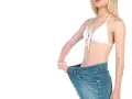 Kobieta w białym bikini pokazuje, jak bardzo schudła, trzymając za luźne dżinsy. Pokonany efekt plateau.
