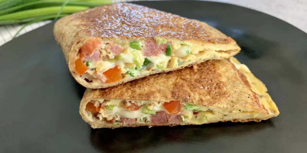 Keto tortilla przepis: złocisty placek z jajek, szynki, pomidorów i szczypiorku, złożony na pół, na czarnym talerzu.