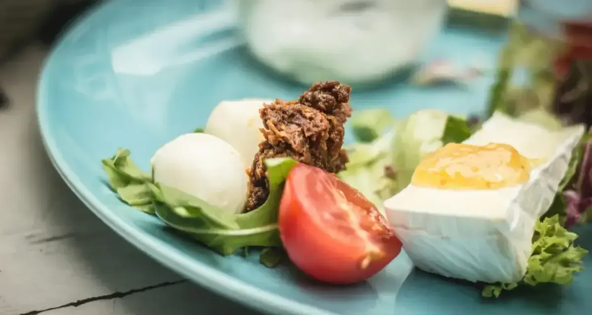 Pyszne keto śniadanie: ser brie z dżemem, kulki mozzarelli, pomidor i rukola na błękitnym talerzu.