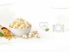 Miseczka popcornu, obok łopatka z ziarnami kukurydzy. Czy popcorn jest lekkostrawny? Ikony serca i talerza sugerują zdrowy posiłek.
