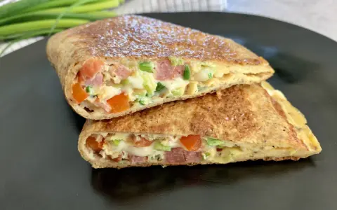 Keto tortilla przepis: złocisty placek z jajek, szynki, pomidorów i szczypiorku, złożony na pół, na czarnym talerzu.