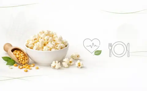 Miseczka popcornu, obok łopatka z ziarnami kukurydzy. Czy popcorn jest lekkostrawny? Ikony serca i talerza sugerują zdrowy posiłek.
