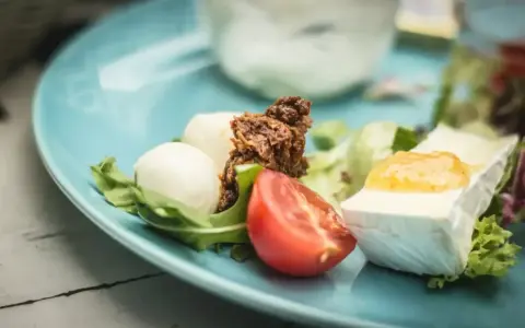 Pyszne keto śniadanie: ser brie z dżemem, kulki mozzarelli, pomidor i rukola na błękitnym talerzu.