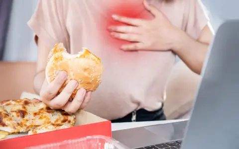 Kobieta z bólem w klatce piersiowej trzyma hamburgera. Zastanawia się, co pić na zgagę po zjedzeniu pizzy i fast foodów.