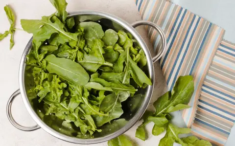 Rukola w durszlaku, idealna do przygotowania jadłospisu tygodniowego dieta FODMAP. Świeże liście na tle kolorowych tkanin.