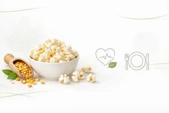 Miseczka popcornu, obok łopatka z ziarnami kukurydzy. Czy popcorn jest lekkostrawny? Ikony serca i talerza sugerują zdrowy posiłek.