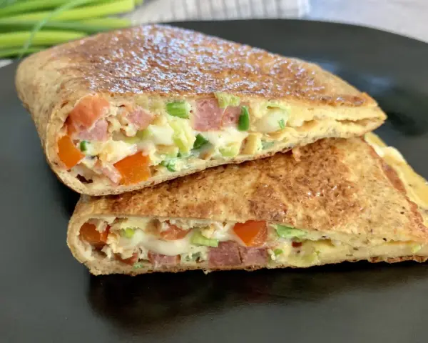 Keto tortilla przepis: złocisty placek z jajek, szynki, pomidorów i szczypiorku, złożony na pół, na czarnym talerzu.