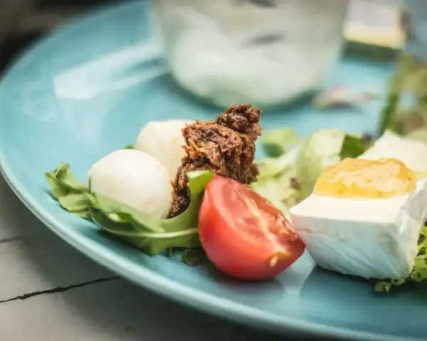 Pyszne keto śniadanie: ser brie z dżemem, kulki mozzarelli, pomidor i rukola na błękitnym talerzu.