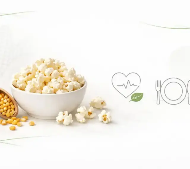 Miseczka popcornu, obok łopatka z ziarnami kukurydzy. Czy popcorn jest lekkostrawny? Ikony serca i talerza sugerują zdrowy posiłek.