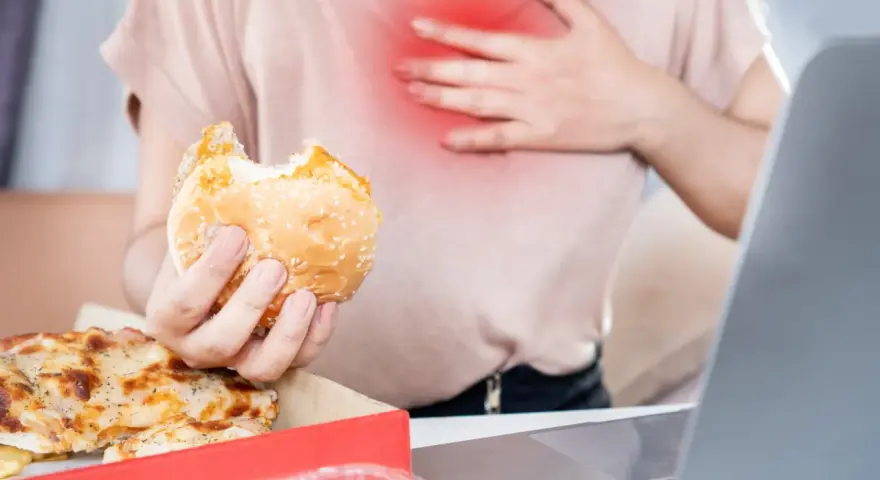 Kobieta z bólem w klatce piersiowej trzyma hamburgera. Zastanawia się, co pić na zgagę po zjedzeniu pizzy i fast foodów.