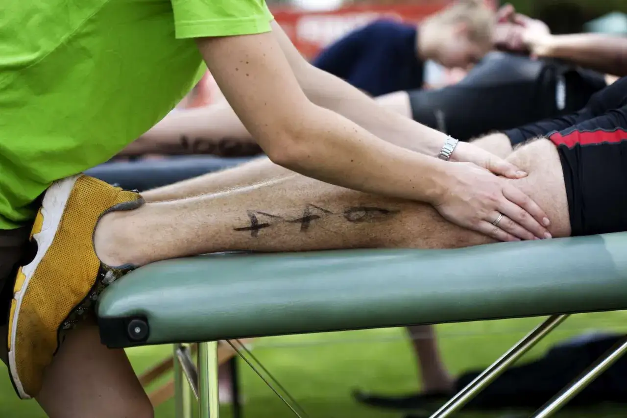 Masażysta rozmasowuje nogę sportowca z widocznymi numerami na łydce, by złagodzić zakwasy po wysiłku.